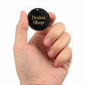GPS Pet Tracker
A hand holding a small round black tag with gold lettering that reads 'Dodos Shop' against a plain white background.