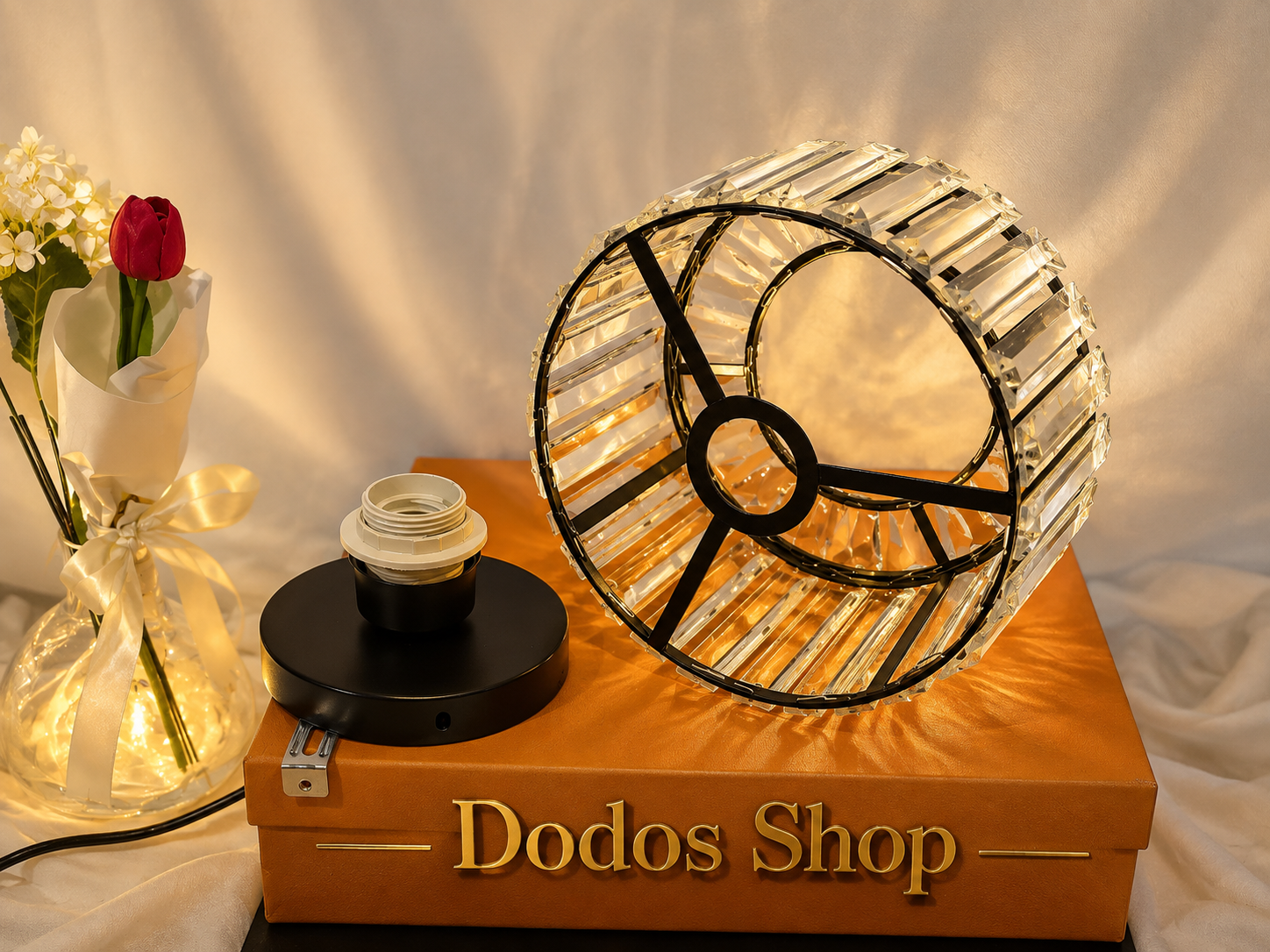 Luxury Crystal Lamp
A clear crystal or acrylic cage-style table lamp with black metal framing, displayed on top of an orange box labeled 'Dodos Shop.' A black lamp base and a vase of flowers sit beside it against a draped white fabric background.