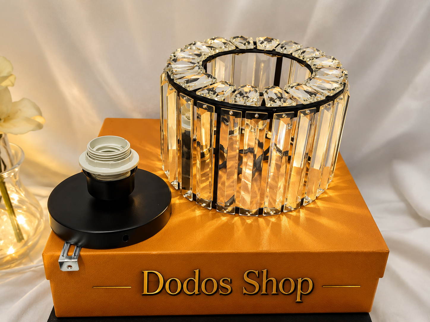 Luxury Crystal Lamp
A clear faceted crystal ring candle holder with a black metal frame, displayed on top of an orange box labeled 'Dodos Shop'. A black socket cover is shown beside it on the box.